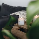 small lit bloom candle on a rustic side table with a dark sofa and grey blanket with a green plant in the foreground