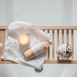 Bottle of bath salts, candle, and towel on a bathtub rim with a neutral background from hobo and co