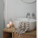 a lit bloom candle and bath salt sby hobo and co in a bathroom setting next to a washbasin and towel