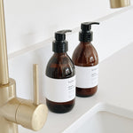 Two amber glass pump bottles with black dispensers and white labels sit on a white sink counter beside a brushed gold faucet. The labels read 'hobo + co 