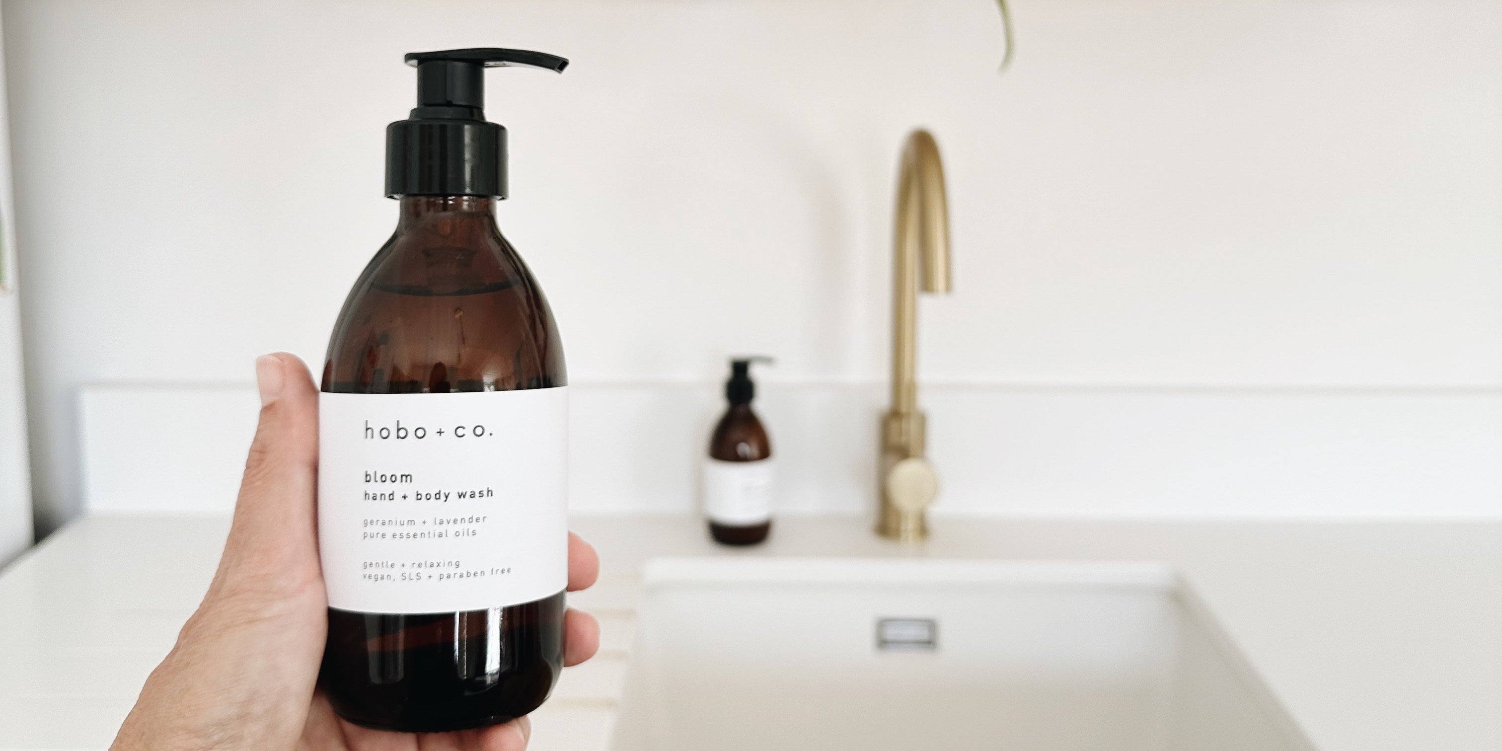 A hand holds a dark amber bottle of hobo co. hand and body wash in front of a bright kitchen sink with wooden shelves and plants in the background.