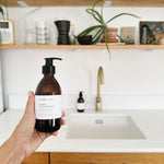 A hand holds a brown amber bottle of hobo + co. hand and body wash in front of a bright kitchen sink with wooden shelves and plants in the background.
