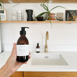 A hand holds a brown amber bottle of hobo + co. hand and body wash in front of a bright kitchen sink with wooden shelves and plants in the background.