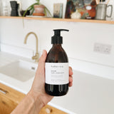 A hand holds a dark amber pump bottle of hobo + co. Bloom hand and body wash in front of a bright, modern kitchen sink with wooden shelves and plants in the background.