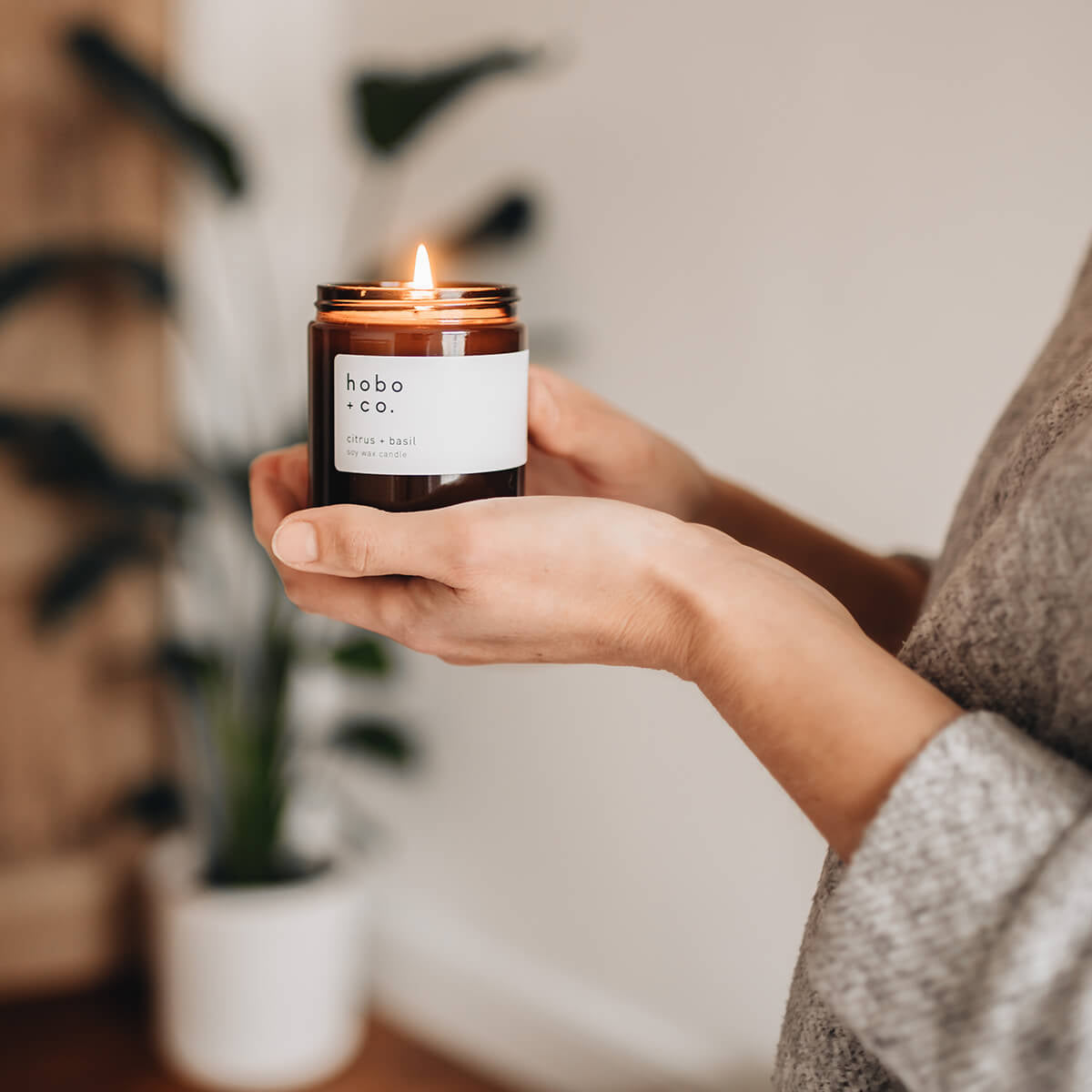 A lit glass jar candle from Hobo and Co held in hands with a plant behind