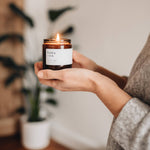 A lit glass jar candle from Hobo and Co held in hands with a plant behind