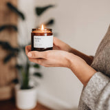 A lit glass jar candle from Hobo and Co held in hands with a plant behind