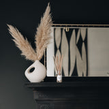 reed diffuser on a black fireplace with a mirror in the background with a cream vase and pampas grass