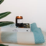 a lit fig medium candle in a brown jar with  flame on top of a book and blanket on a white background
