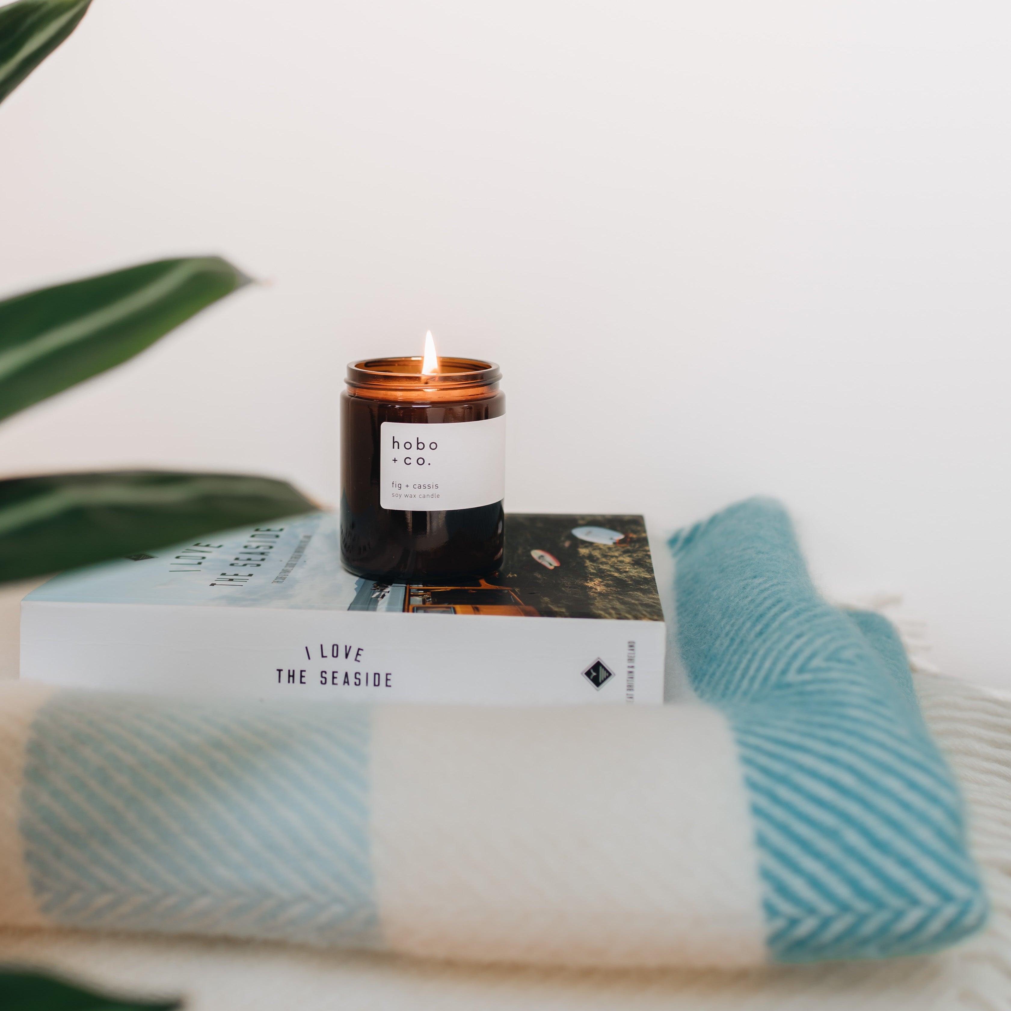 a lit fig medium candle in a brown jar with  flame on top of a book and blanket on a white background