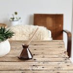 glass diffucer bottle on a rustic table with a renn plant and a brown chair 