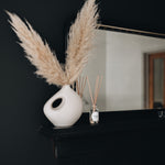 White vase with pampas grass on a black surface with a mirror in the background with reed diffuser bottle on a mantlepiece