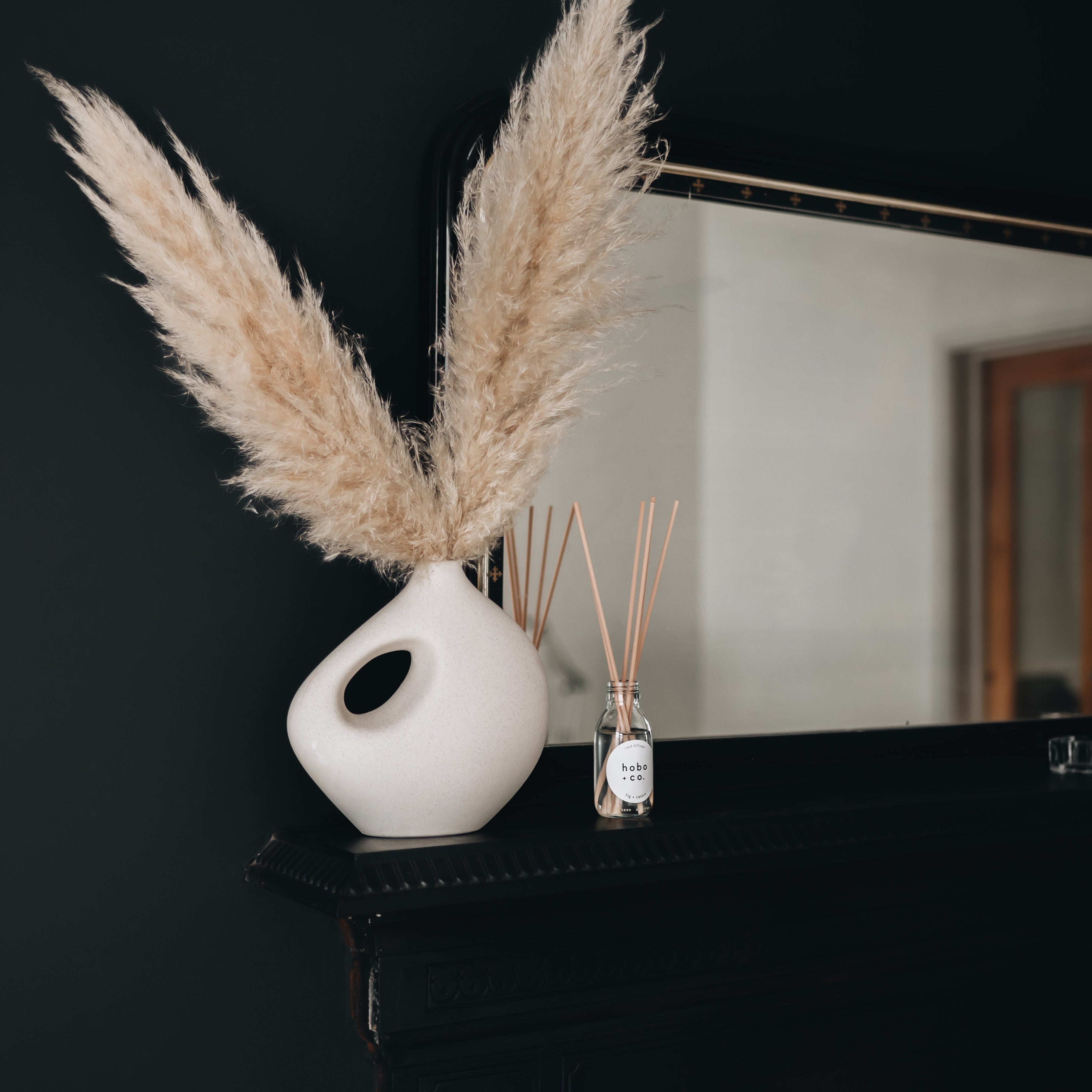 White vase with pampas grass on a black surface with a mirror in the background with reed diffuser bottle on a mantlepiece