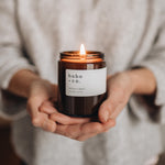 Person holding a lit citrus and basil candle labeled 'hobo & co.'