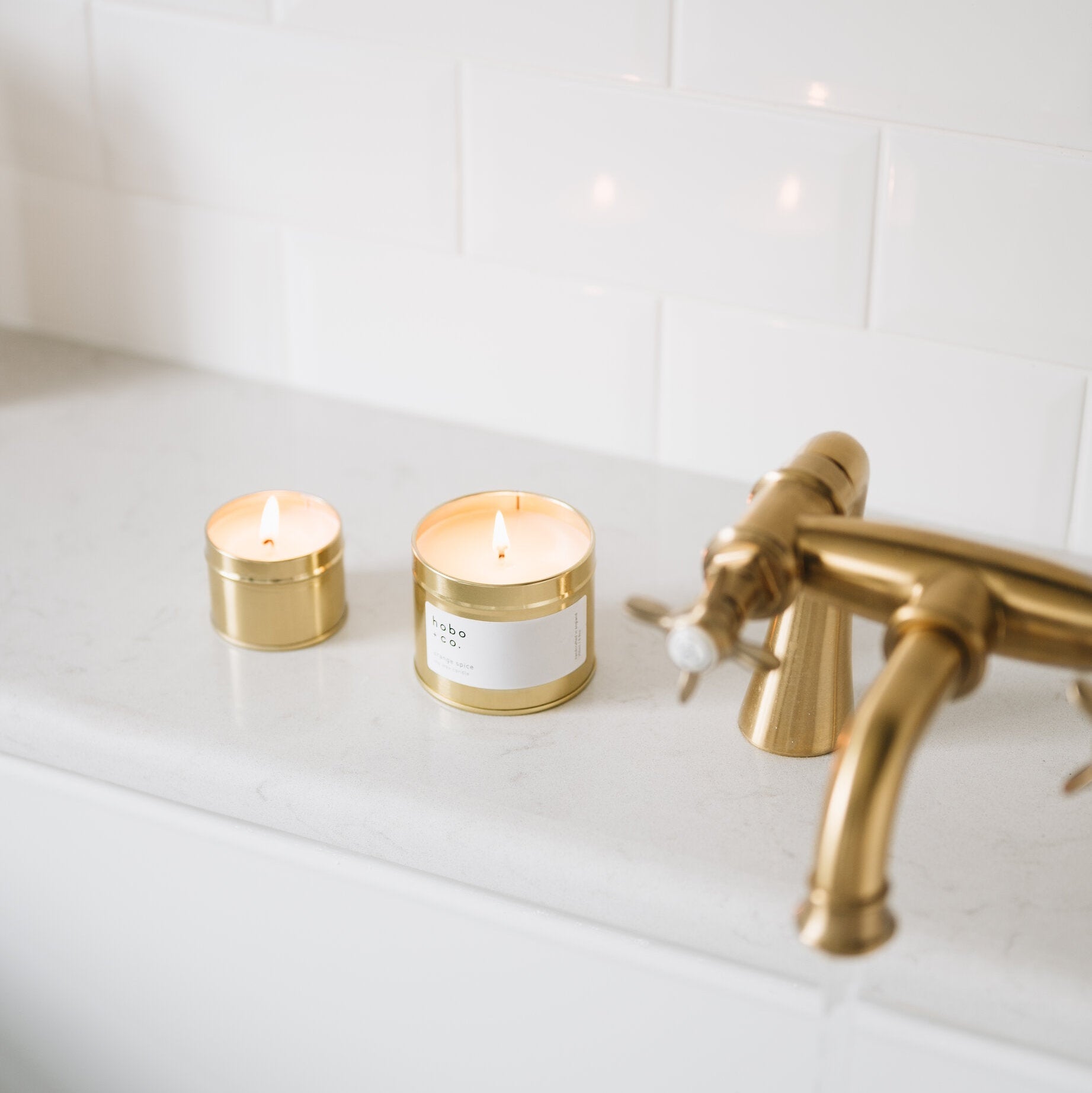 Two candles on a bathtub rim with a gold faucet against a white tiled wall.