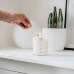 large candle being lit by a match held in a hand on a shelf