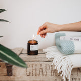 Hand lighting a candle on a wooden surface with a white wall and plant in the background