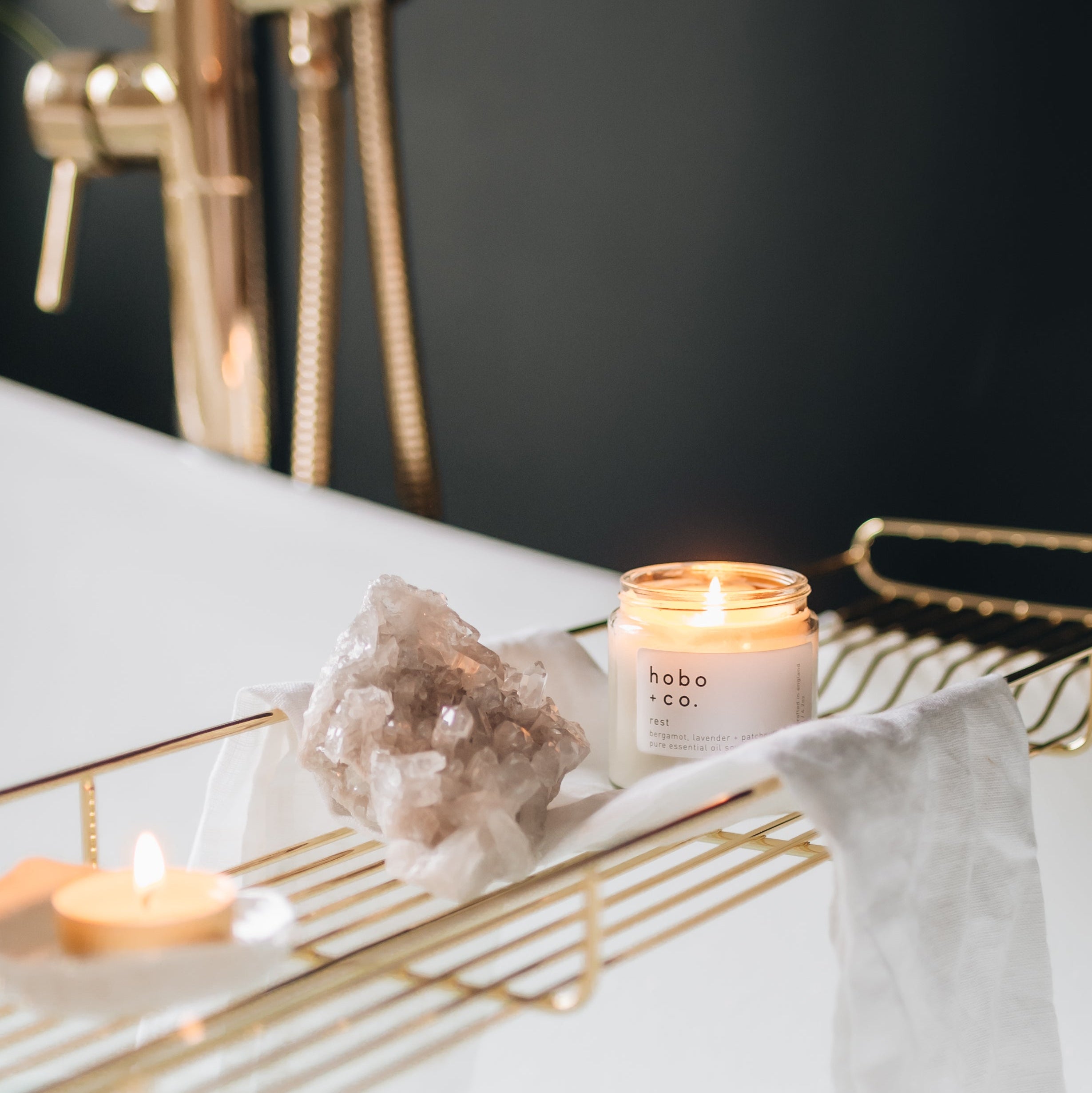 Lit candle with 'hobo' branding on a gold bath tray on a bath with a dark background