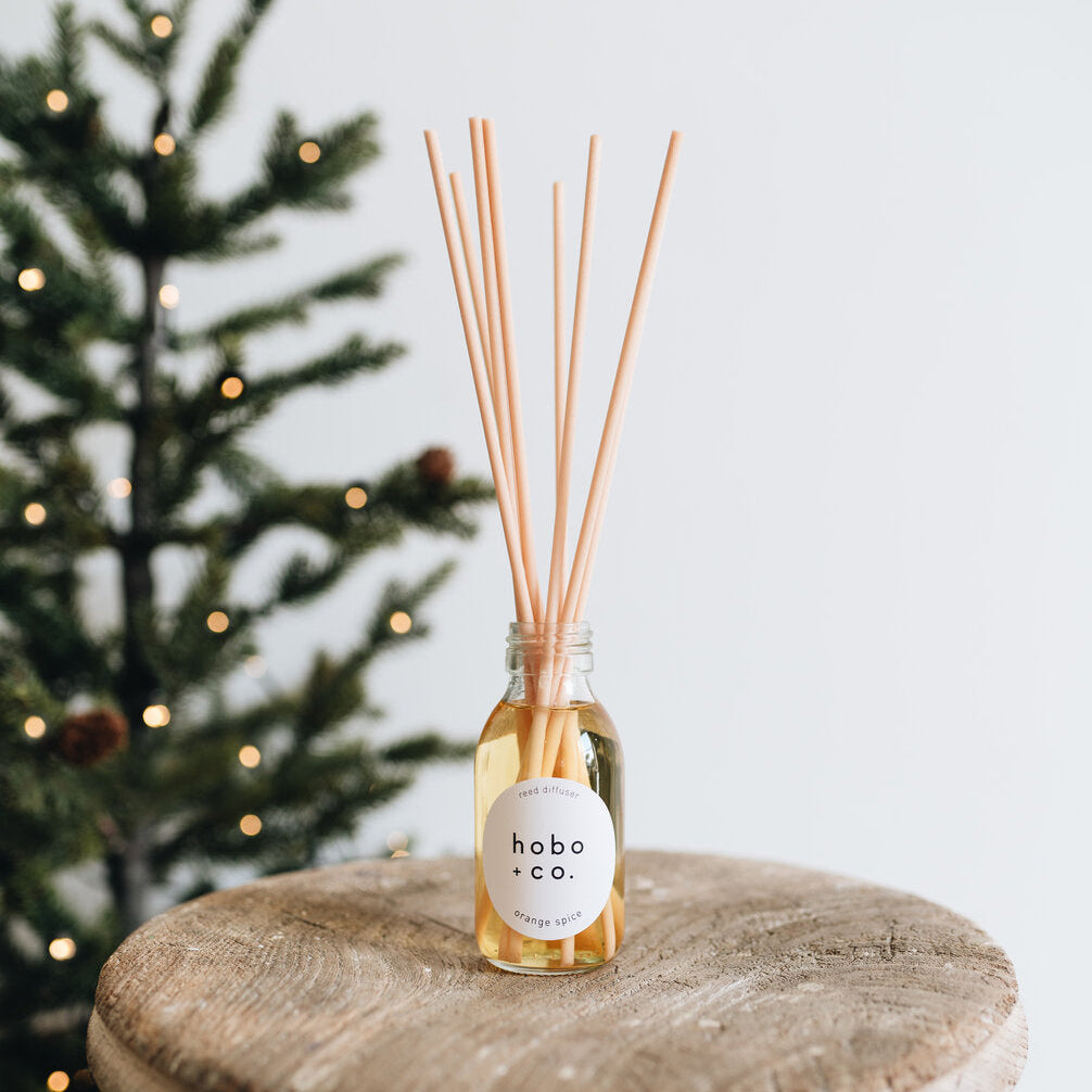 Reed diffuser with 'hobo + co' label on a wooden surface with a blurred Christmas tree in the background.