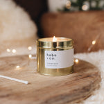 Lit orange spice candle in a large gold tin in a cosy living room setting with sheepskin rug and fairy lights