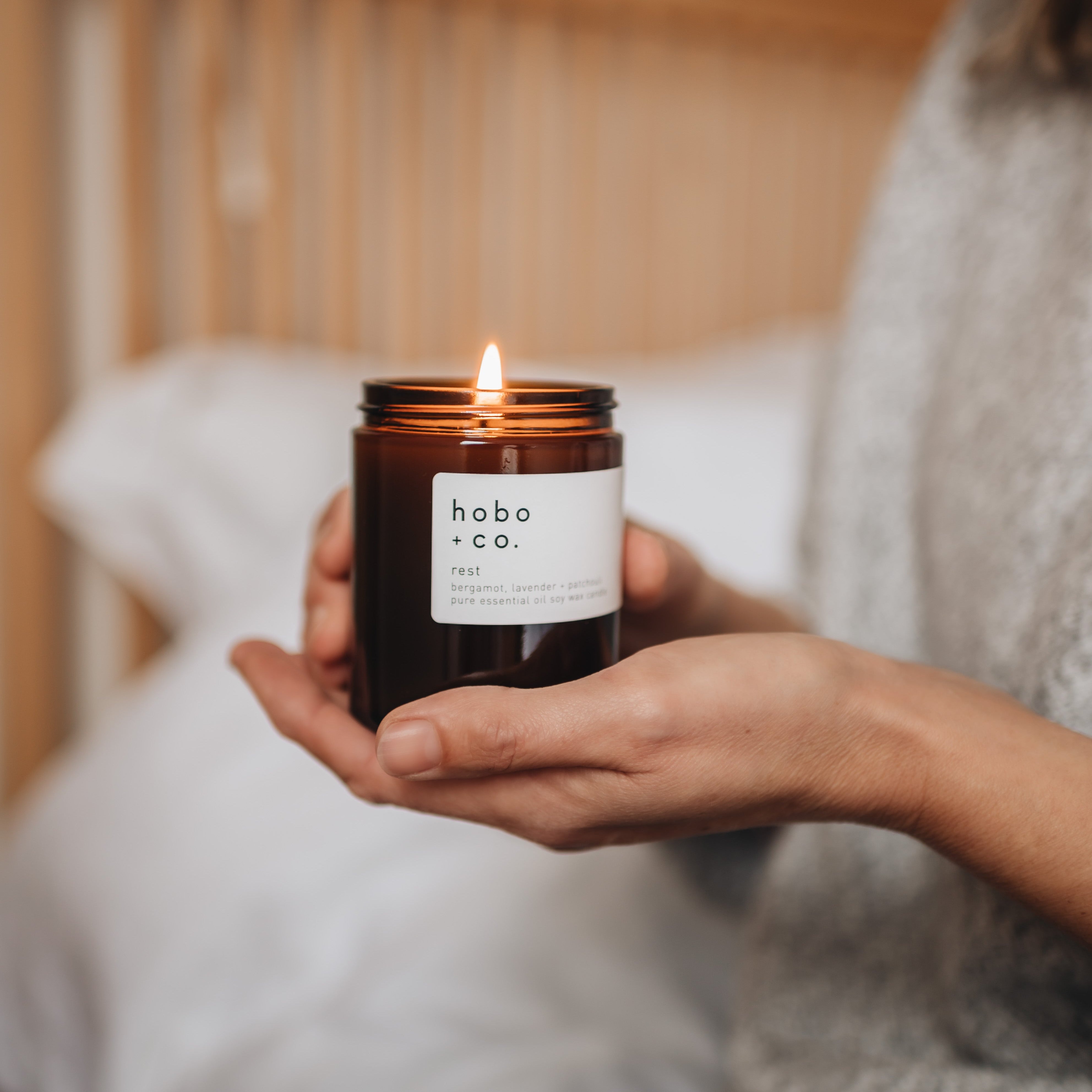 Person holding a lit candle with 'hobo & co.' label in a cozy indoor setting