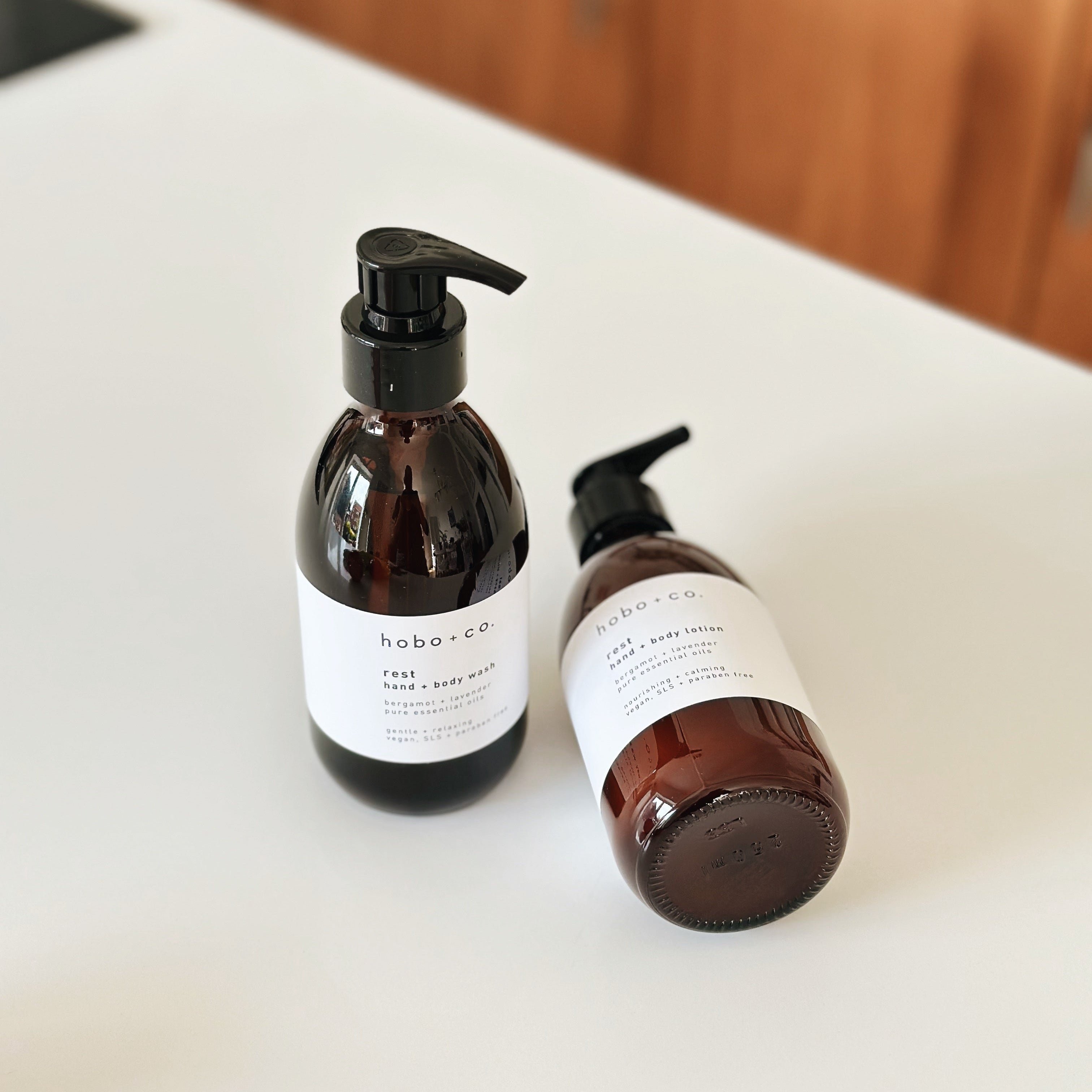 Two amber glass pump bottles with black dispensers and white labels from the brand hobo + co, placed on a white countertop with a blurred wooden background.
