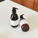 Two amber glass pump bottles with black dispensers and white labels from the brand hobo + co, placed on a white countertop with a blurred wooden background.
