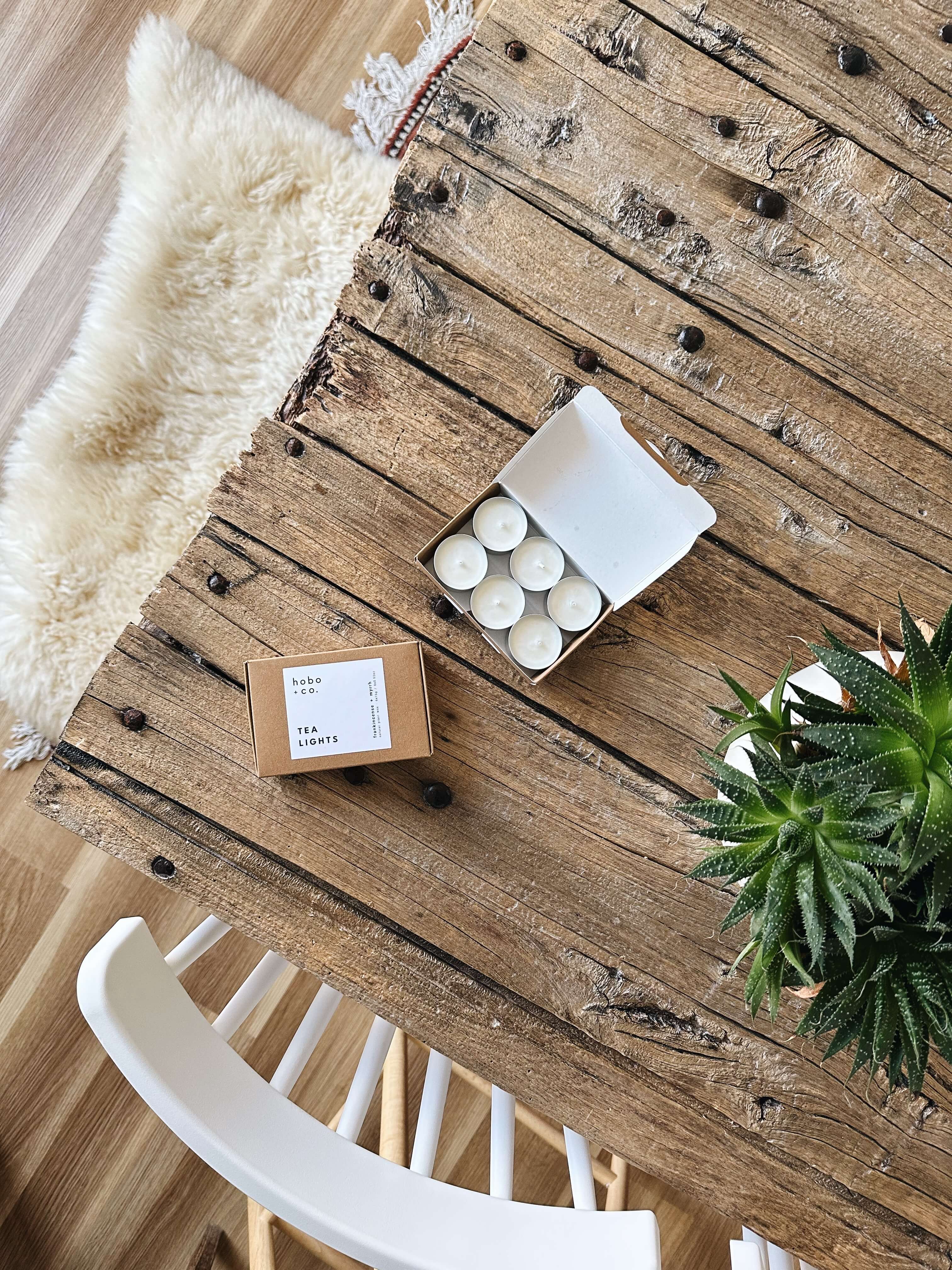 Rustic wooden table with a box of white festive tealights and a plant on a wooden floor with a cosy sheepskin rug.