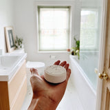 Hand holding a white shower steamer in a bathroom with a bath, shower and sink. Citrus aromatherapy shower steamer for self care