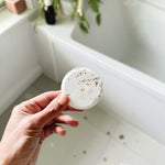 Hand holding a white shower steamer over a bathtub with plants in the background. Natural essential oil wellness shower steamer for morning routine