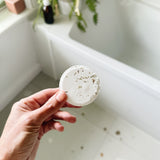 Hand holding a white shower steamer over a bathtub with plants in the background. Natural essential oil wellness shower steamer for morning routine