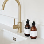 A matte brass single-handle kitchen faucet installed above a white sink, with two amber pump bottles and white labels on the countertop against a white tiled wall.