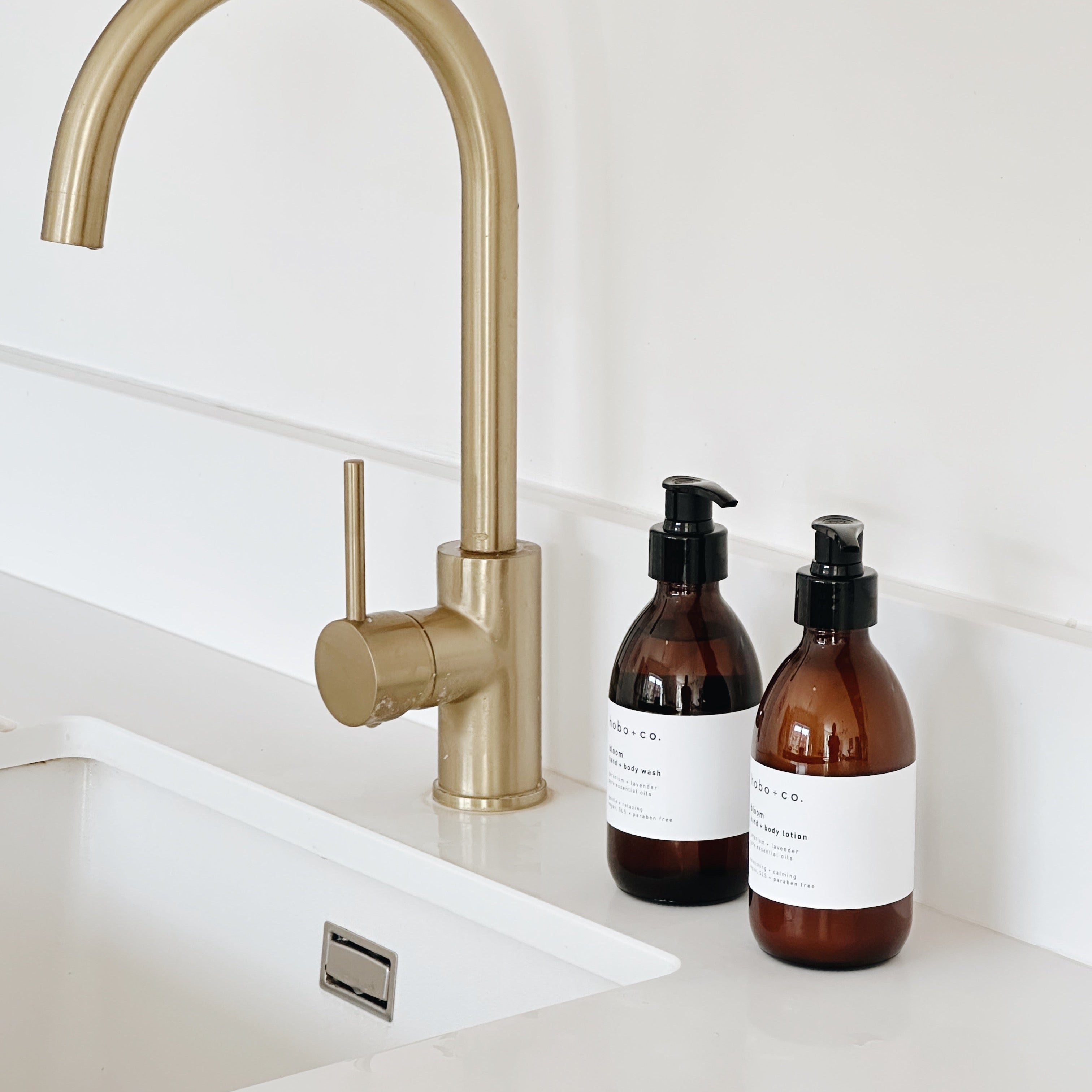 A matte brass single-handle kitchen faucet installed above a white sink, with two amber pump bottles and white labels on the countertop against a white tiled wall.