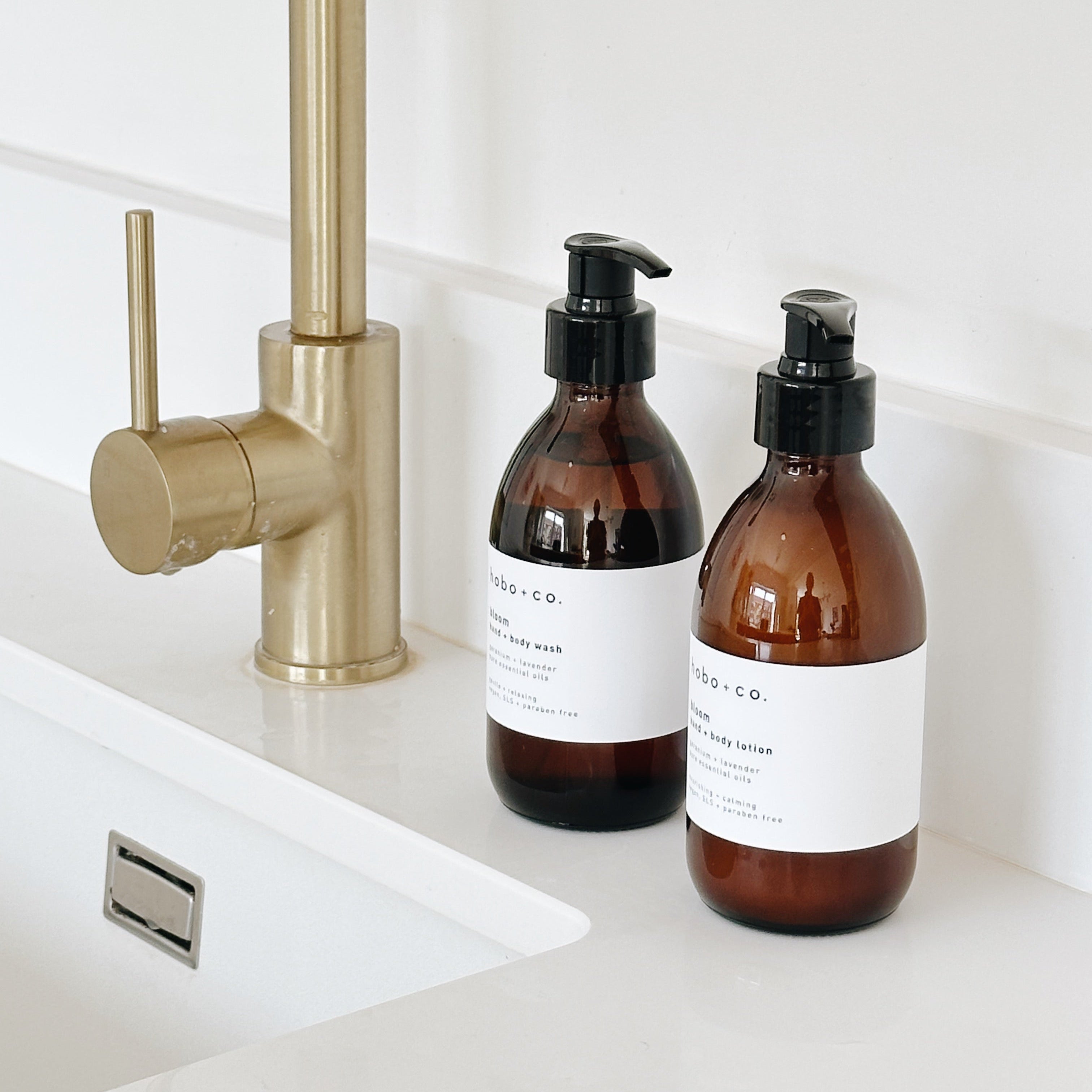 Two amber glass pump bottles with white labels and black dispensers sit on a white sink counter beside a brushed brass faucet.